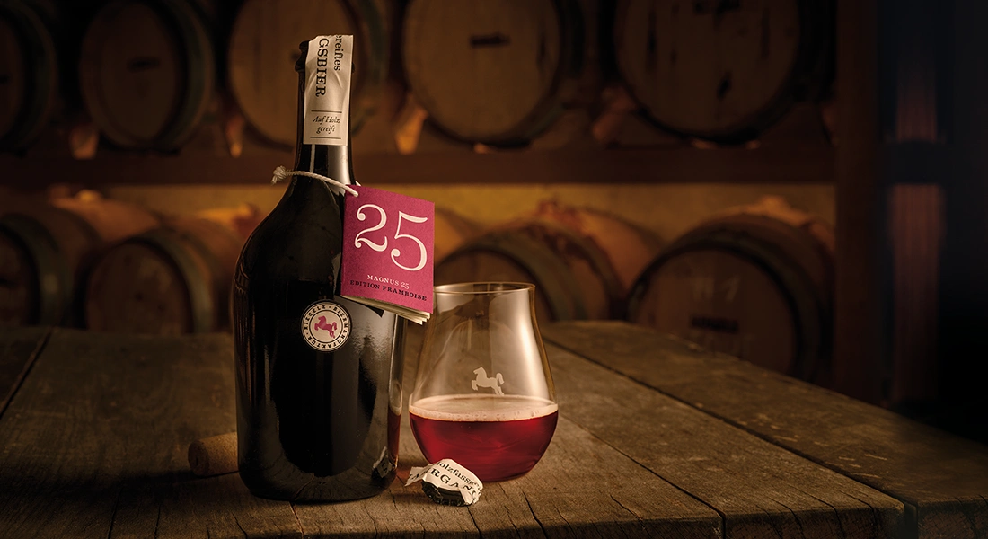Atmosphärisches Foto einer Flasche Riegele Magnus 25 mit Glas vor Holzfässern