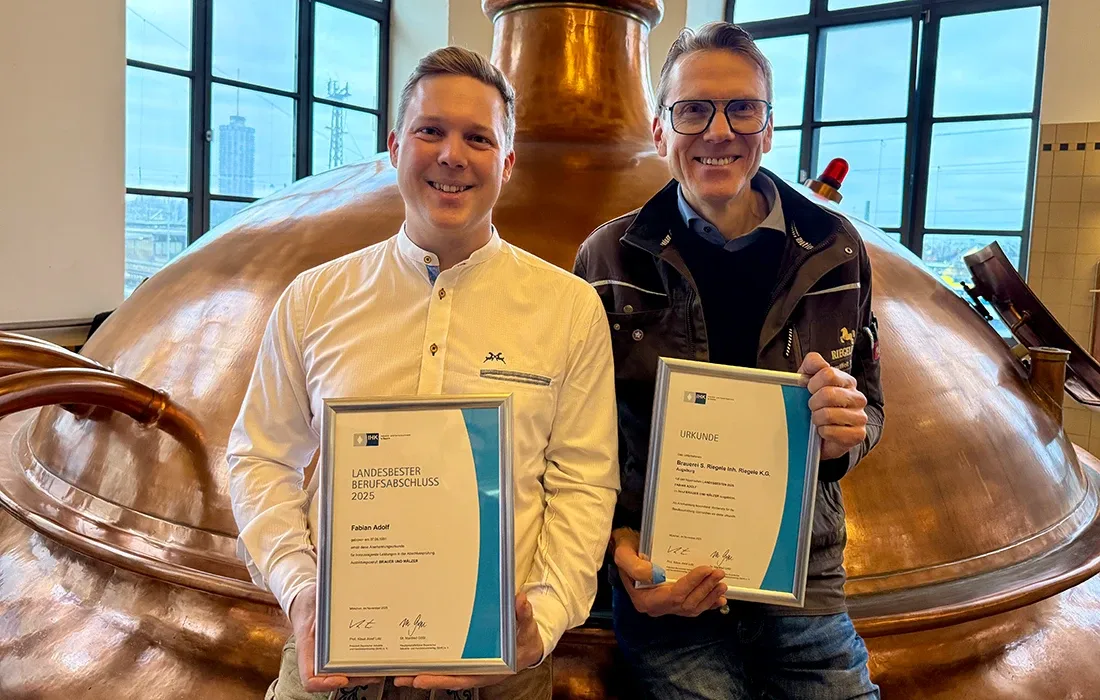 Fabian Adolf und Ausbilder Frank Müller, lachend mit ihren IHK-Urkunden vor einem Sudkessel