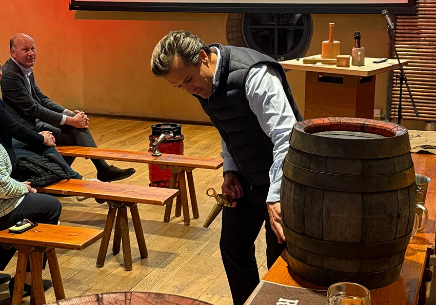 Sebastian Priller neben historischen Holzfass, erklärt wo angezpaft wird und zeigt auf das FAss